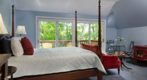 Bedroom with dark wooden four-poster bed, white bedding, red, white, and blue pillows, red upholstered love seat, large patio doors out to balcony