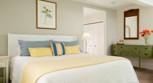 Bedroom with white wooden bed, white bedding, yellow blanket, yellow and blue pillows, and olive buffet table