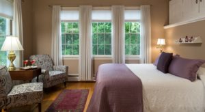 Bedroom with white bedding, purple blankets and pillows, upholstered chairs, and large windows with views of green trees