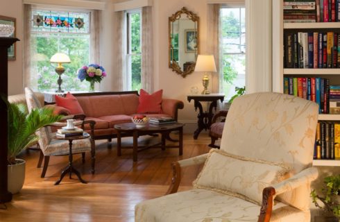Large living room with white walls, hardwood flooring, wooden furniture with multiple-colored upholstery, and large book shelf