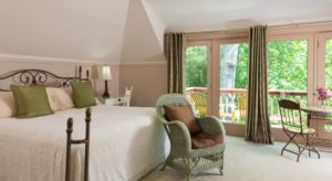 Bedroom with rod-iron bed, white bedding, olive pillows, light green wicker chair with brown cushions, large patio doors leading out to a balcony