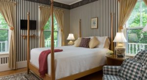 Bedroom with black and white striped wallpaper, light wooden four-poster bed, white bedding, burgundy blanket, and green and white plaid upholstered chair