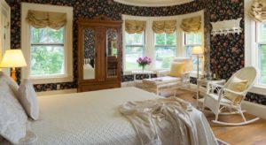 Bedroom with floral wallpaper, white bedding, white wicker furniture, large wooden armoir with mirrors, and many windows