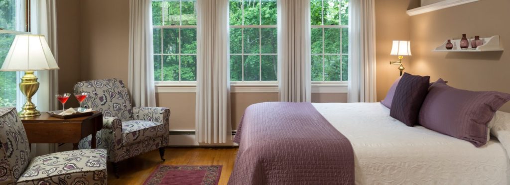Bedroom with white bedding, purple blankets and pillows, upholstered chairs, and large windows with views of green trees