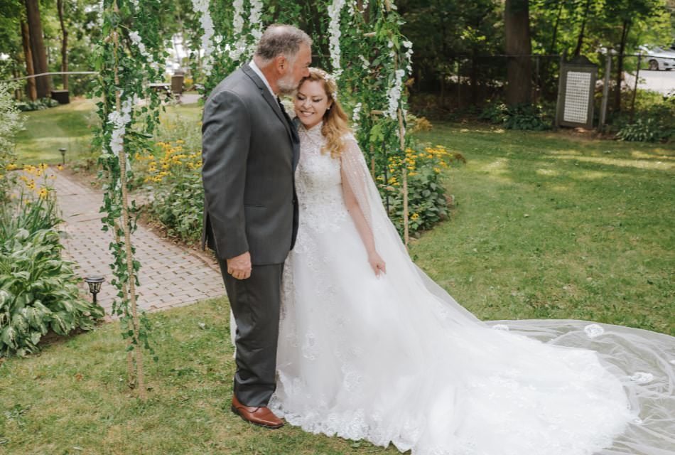 Bride in white dress with man in gray suit kissing the bride's forehead standing in green grass