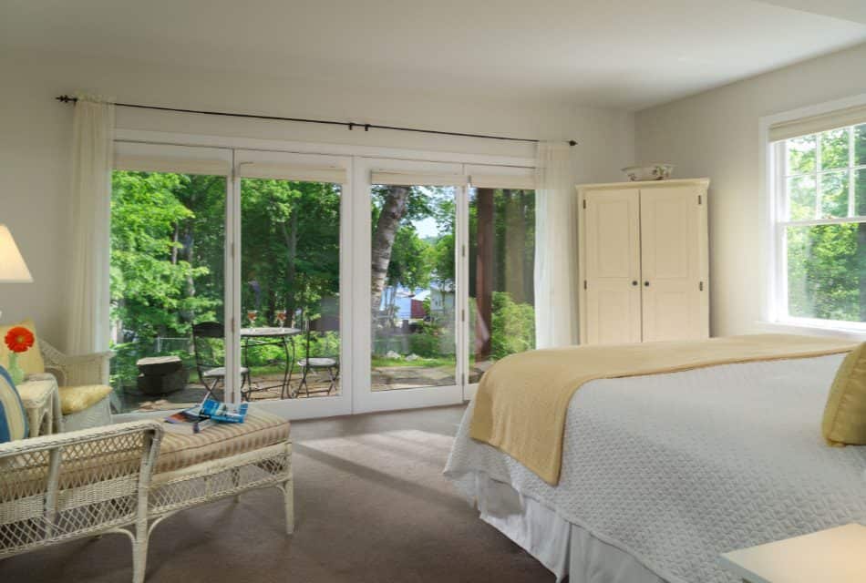 Bedroom with white bedding and yellow blanket, wicker furniture, armoir, and large patio door with a view to the outside patio