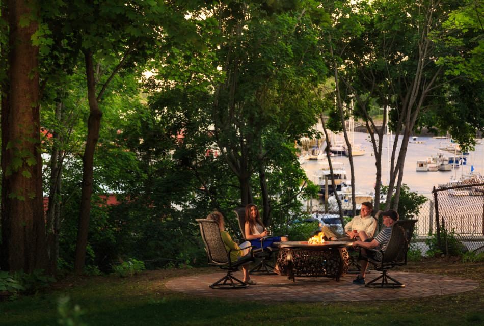 People sitting in patio furniture around a firepit surrounded by large green trees with a view of the harbor in the background
