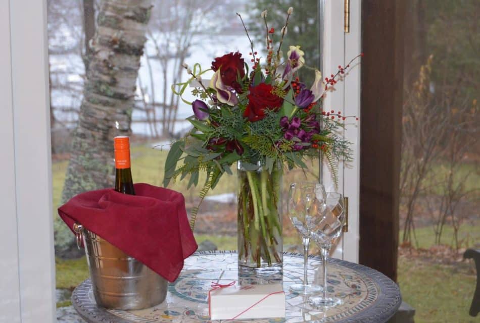 Ice bucket chilling some wine, glass vase with a bouquet of red and purple flowers, two wine glasses, and small white box